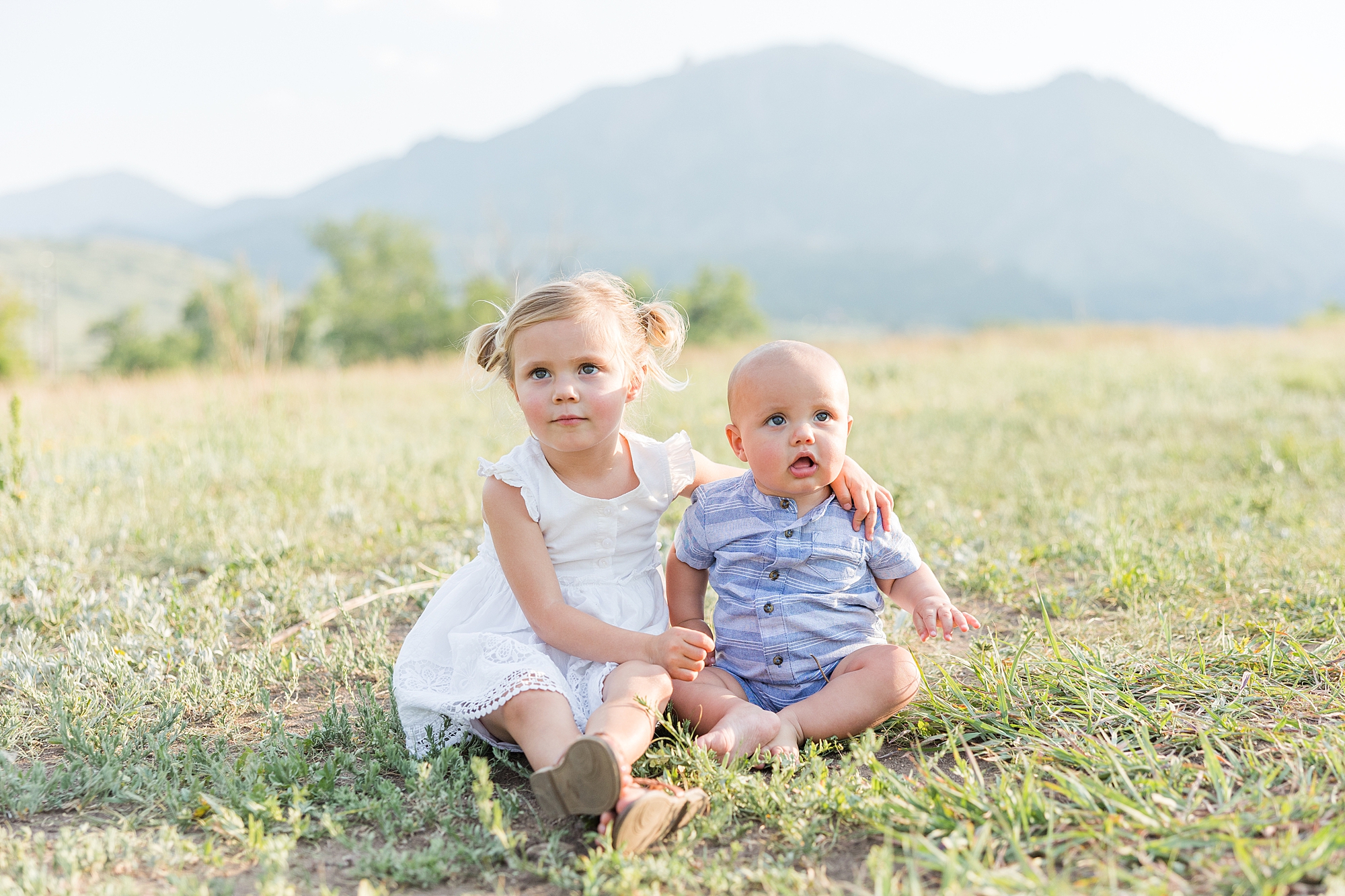 sister sits in grass with arm around baby brother 