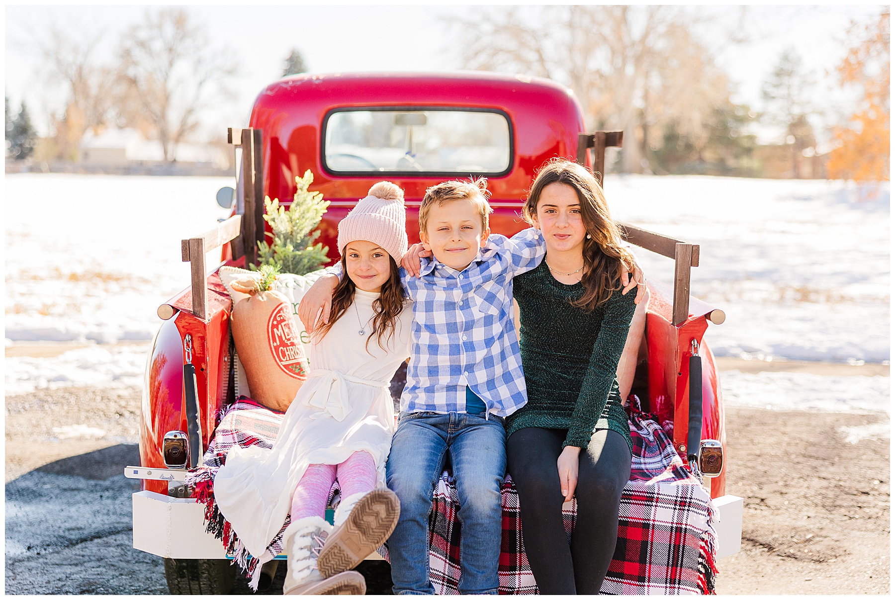 three siblings sit with arms around each other in the back of a red truck