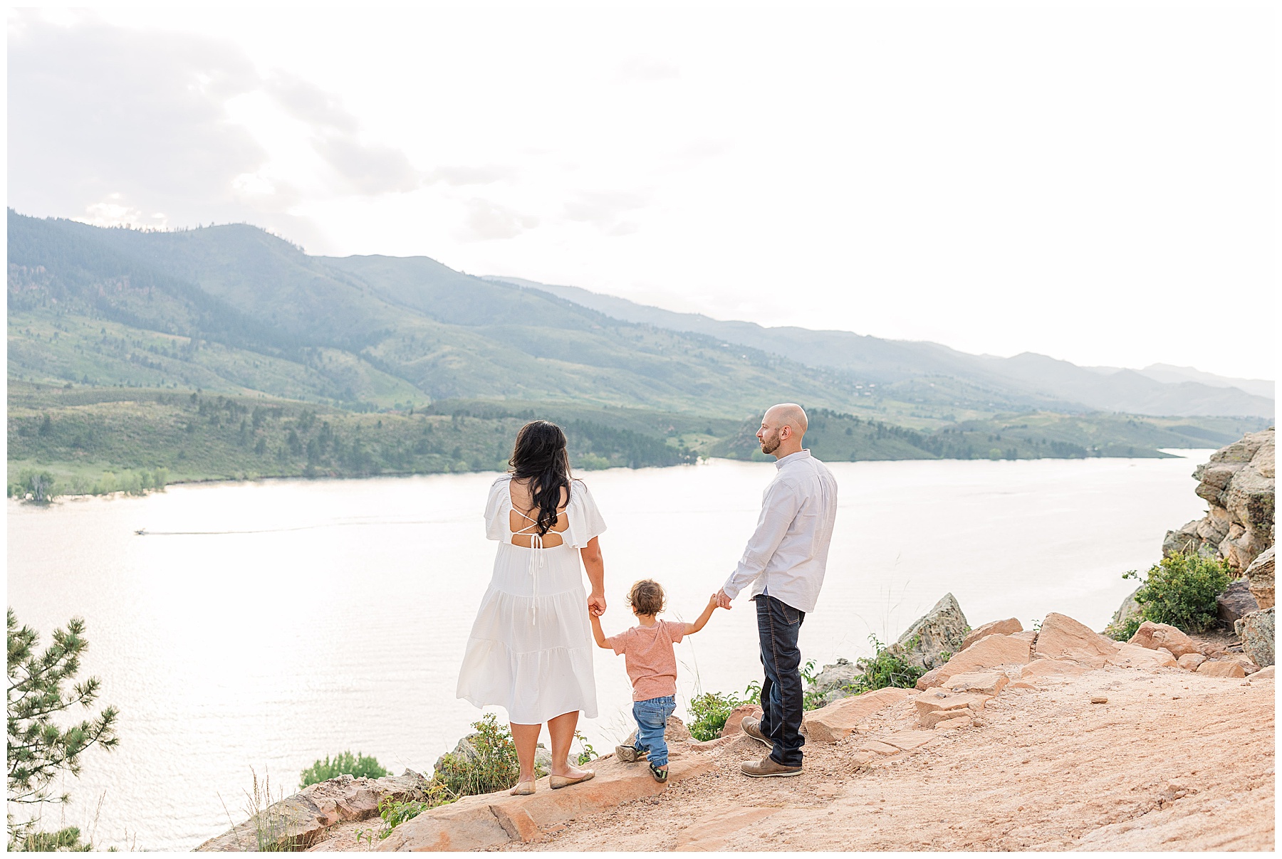 Expecting mother and father hold the hands of their toddler and look out over a river staring off to the mountain behind it
