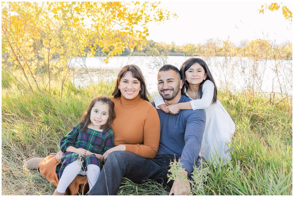 Family of four sit in the tall grass with a lake and golden apens behind them for light and airy outdoor fall photos