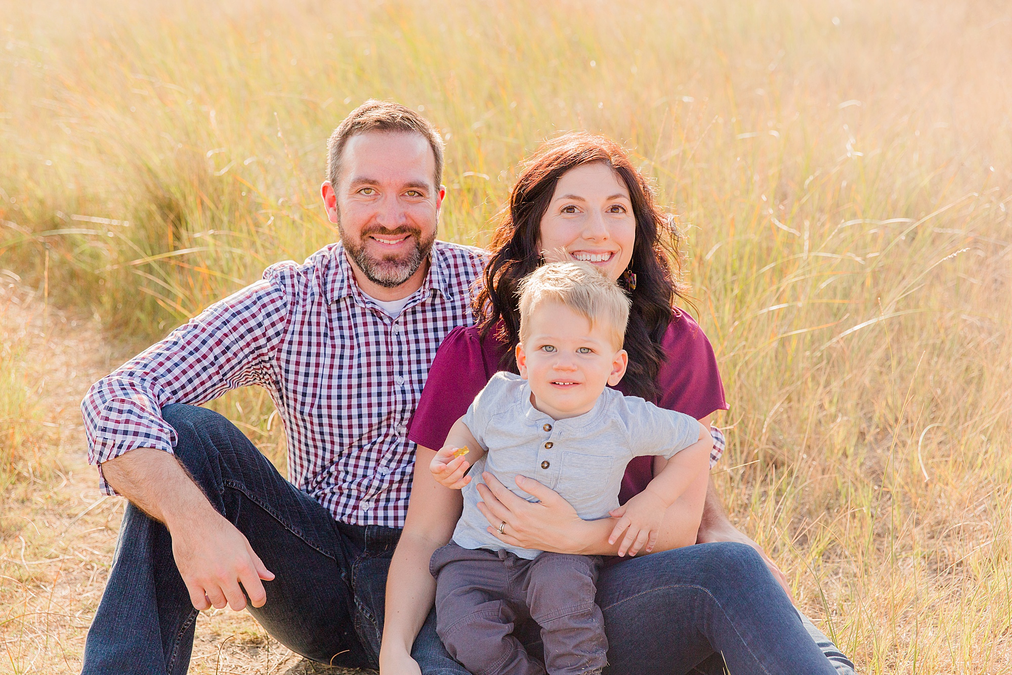 mom and dad sit in tall grass with their son during fall session