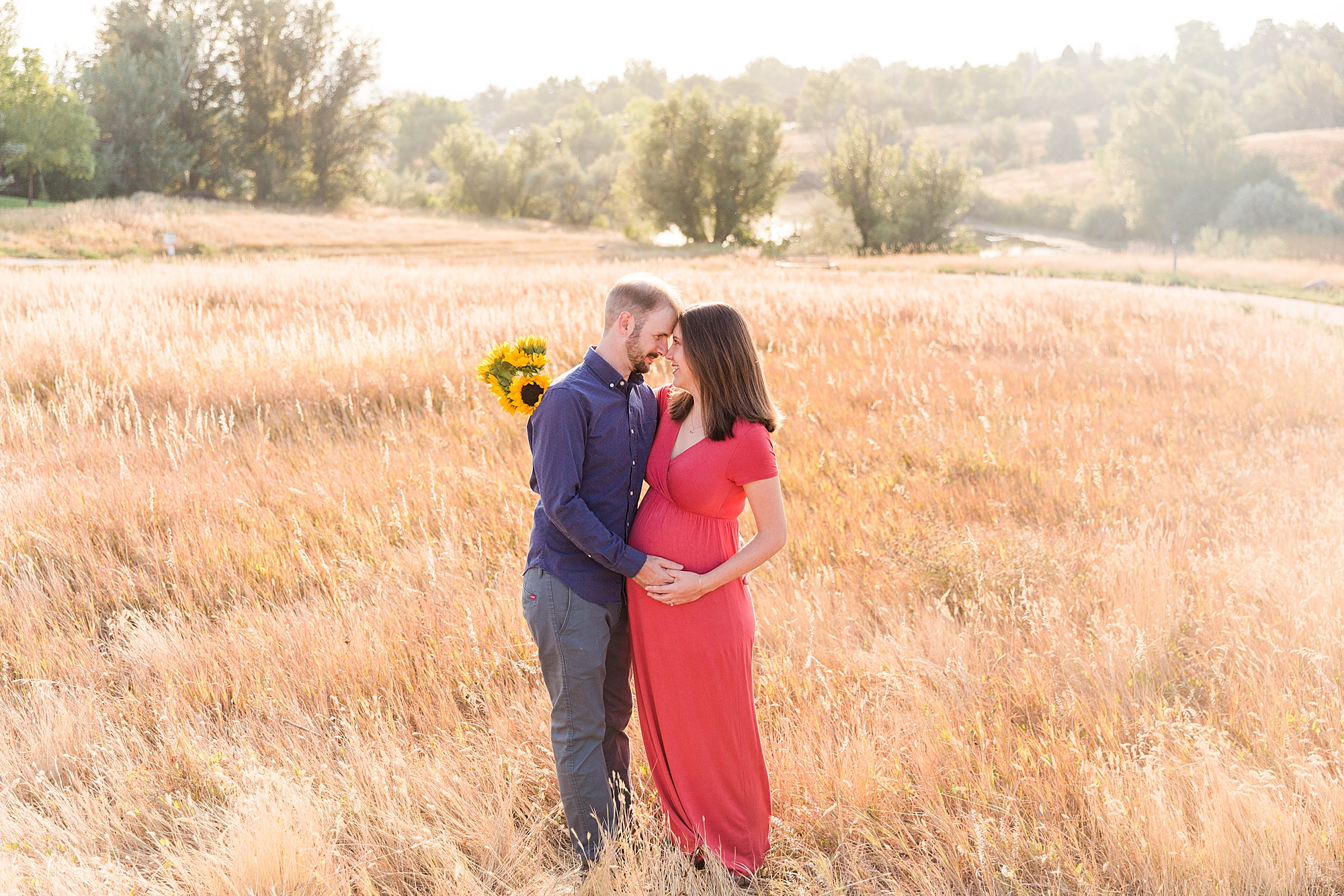 couple hug in field while mom holds sunflowers during maternity shoot