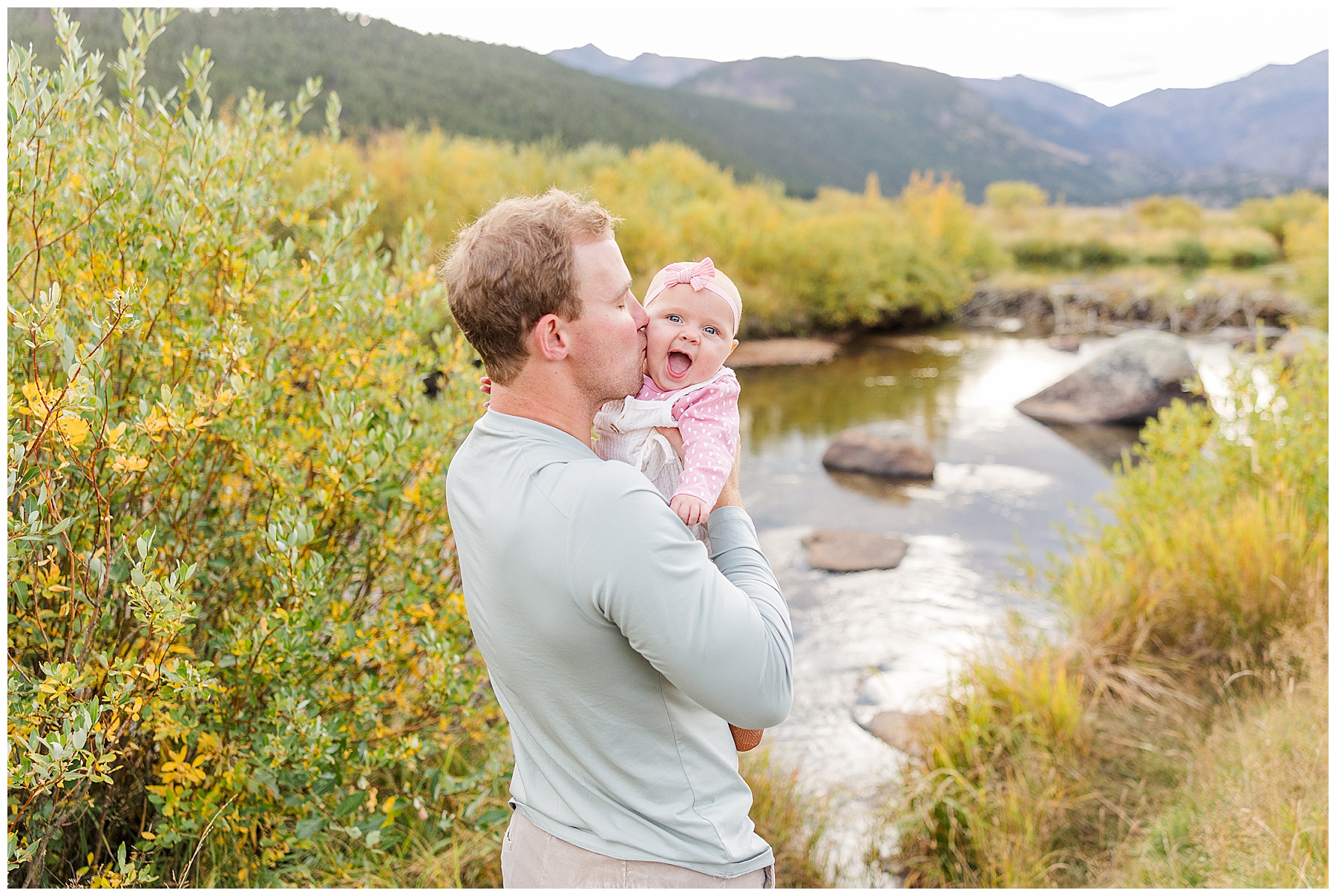 daddy kisses baby girl on the cheek with a pond in the background