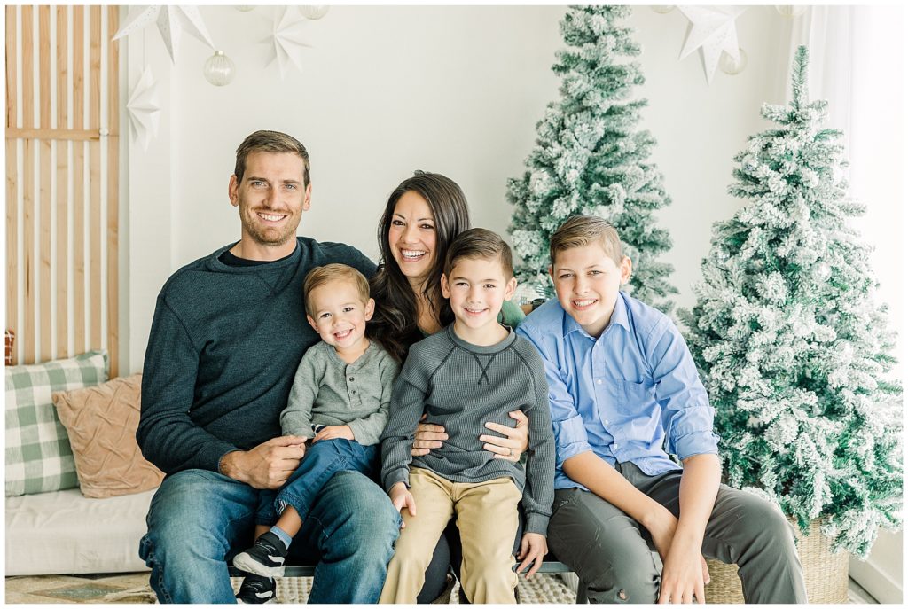family of five pose sitting in front of Christmas trees