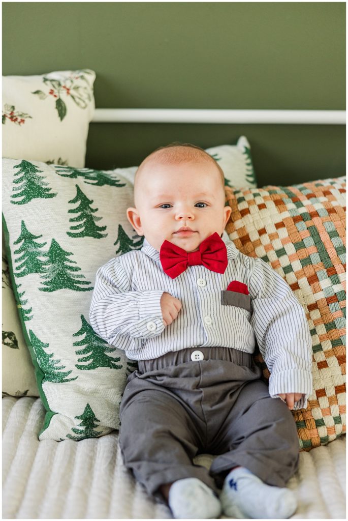 baby in a bright red Christmas bowtie sits in cozy pillows during Christmas minis