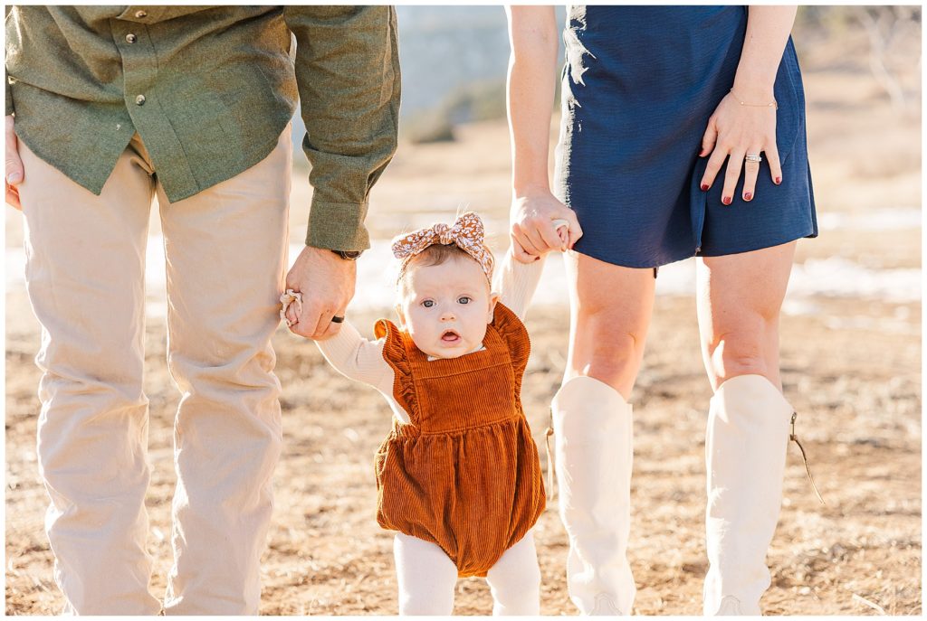 Baby girls stands holding onto mom and dads hands for Cohen Extended Family Session