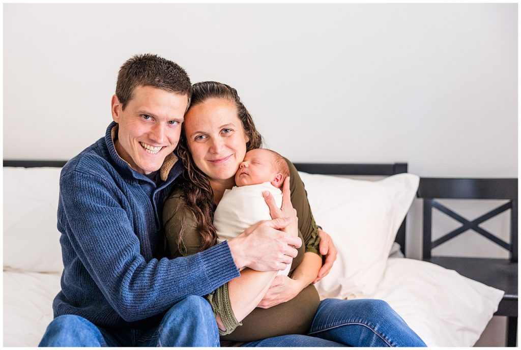 husband and wife sit on bed posing with their baby for a photo session with Catherine Chamberlain Photography