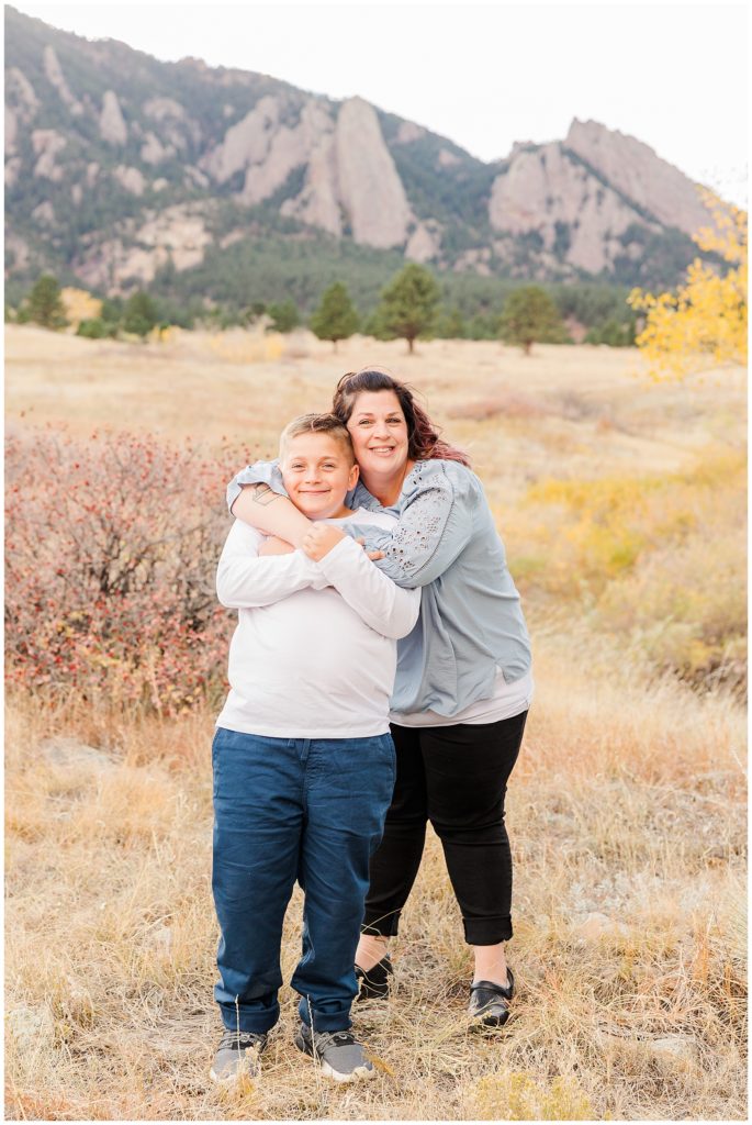 Son grabs his mom's arms which are wrapped around him while posing for family photos in front of the flatirons located in Boulder, Colorado
