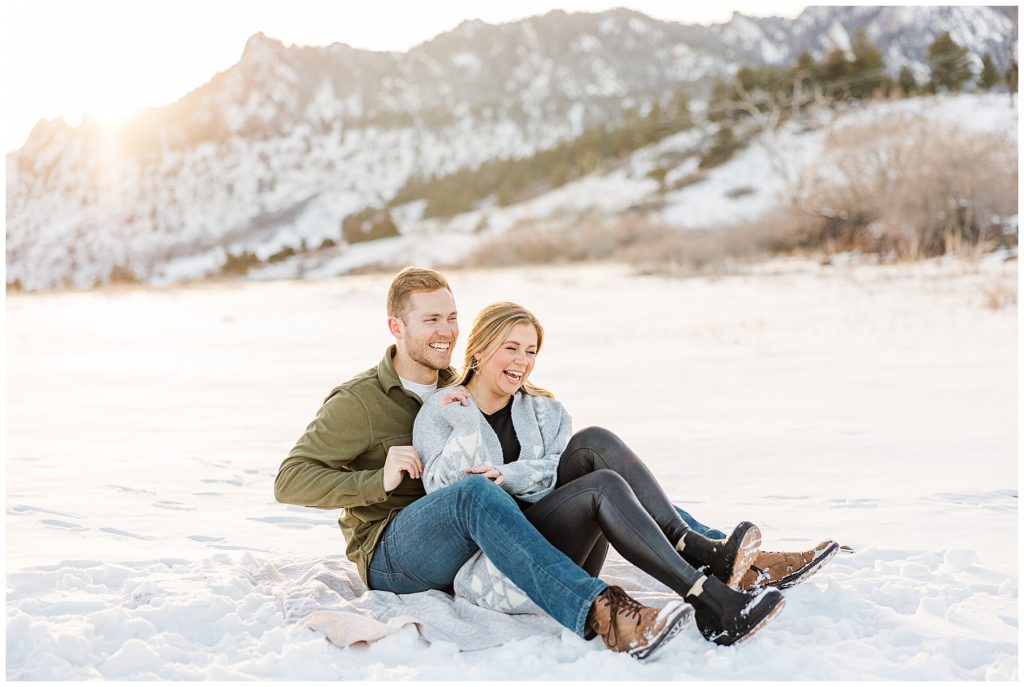 Young couple tickle each other and laugh during a winter photoshoot while seated on the ground. 
