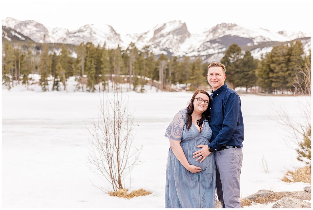 Couple snuggle together and put hands on wife's pregnant belly for their maternity session in the snow