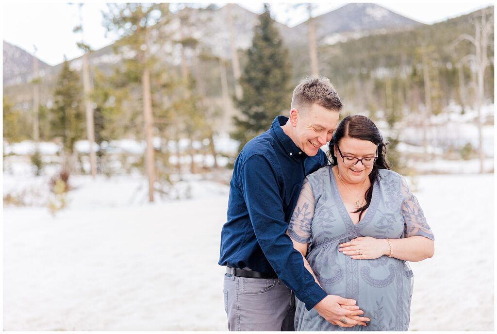 Couple hold hands and look down at her pregnant belly of their first child in Northern Colorado at their photo shoot in Rocky Mountain National Park