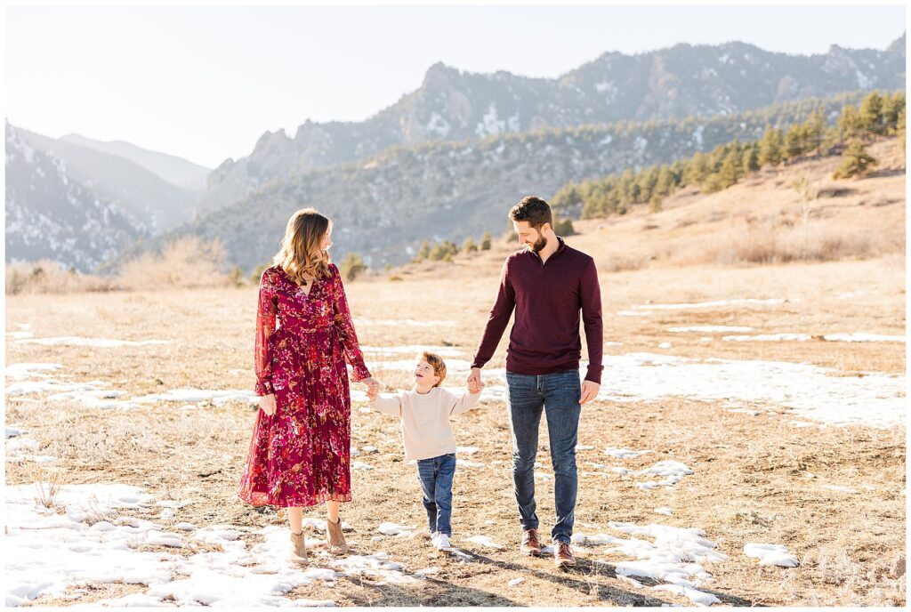 Father and Mother hold their son's hands while walking together in front of a mountain at South Mesa Trailhead in Boulder Colorado