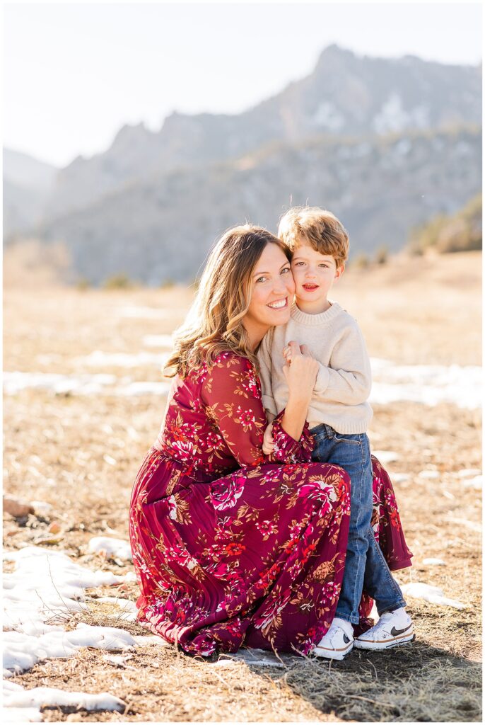 Mother in a beautiful long dress squats down cheek to cheek with her young son and they hold hands and post for the Martin Family Outdoor Session in Boulder Colorado