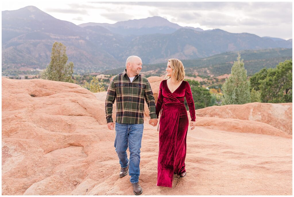 Husband and wife hold hands and look at each other while walking on red rock with a mountainous background in Northern Colorado Outdoor Family Photo