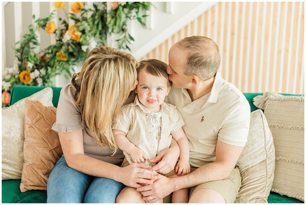 Mom and dad kiss their son's cheek who is seated between them on a neutral colored couch for light and airy photos with Catherine Chamberlain
