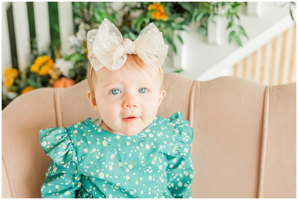 Close up of baby girl in a green romper with a white bow sitting on a neutral colored couch with a floral staircase behind her