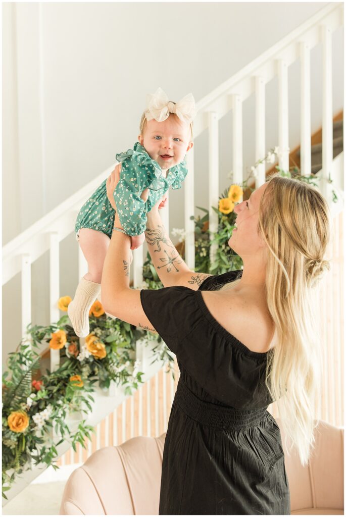Mother holds her baby up in the air in front of a floral decorated staircase at Sugarhill Studio in Longmont, CO