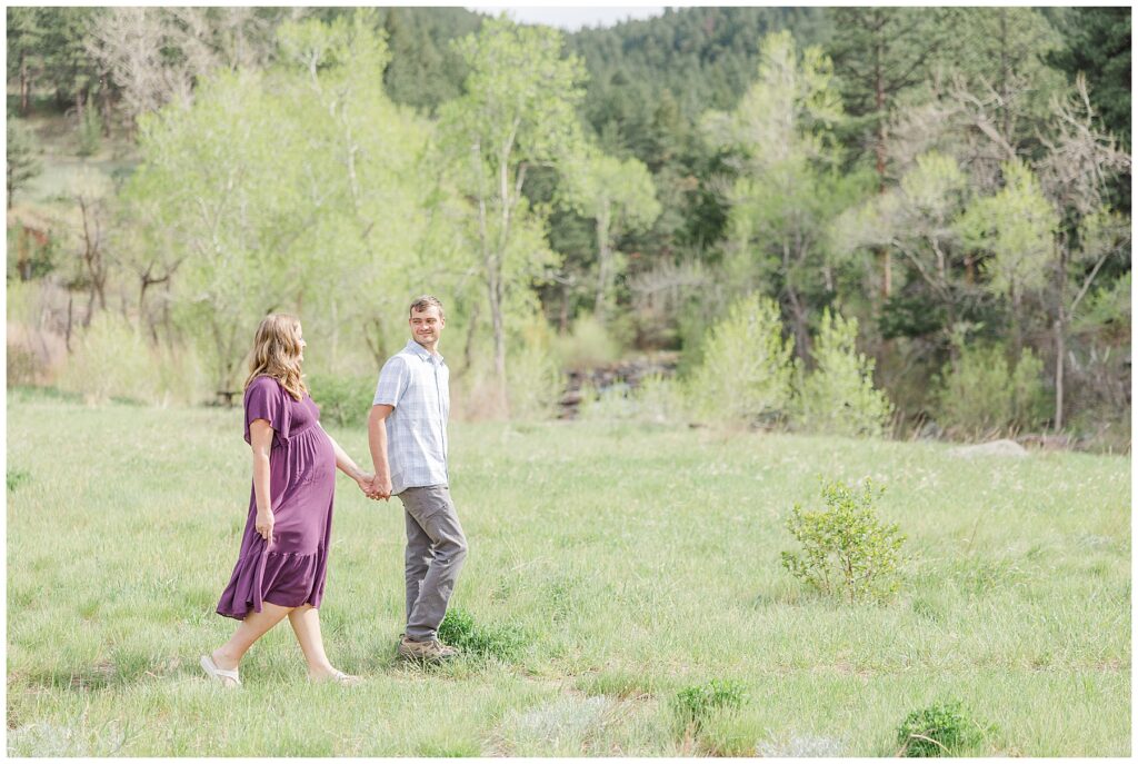 Expecting couple walk in a grassy field holding hands and the husband looks back at his pregnant wife for pictures in Boulder, CO