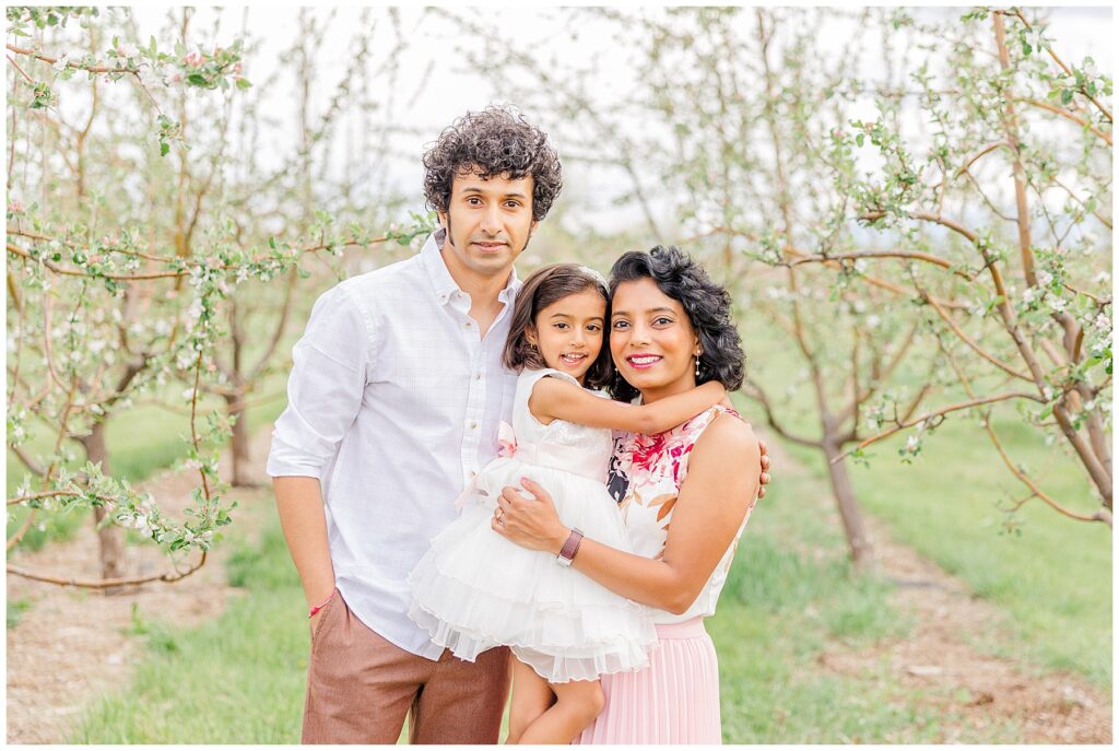 Father, mother, and daughter stand in the middle of an orchard for spring photos 