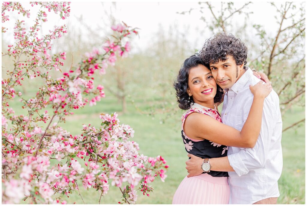 Husband and wife stand sideways hugging each other next to a pink budding tree in Longmont, Colorado