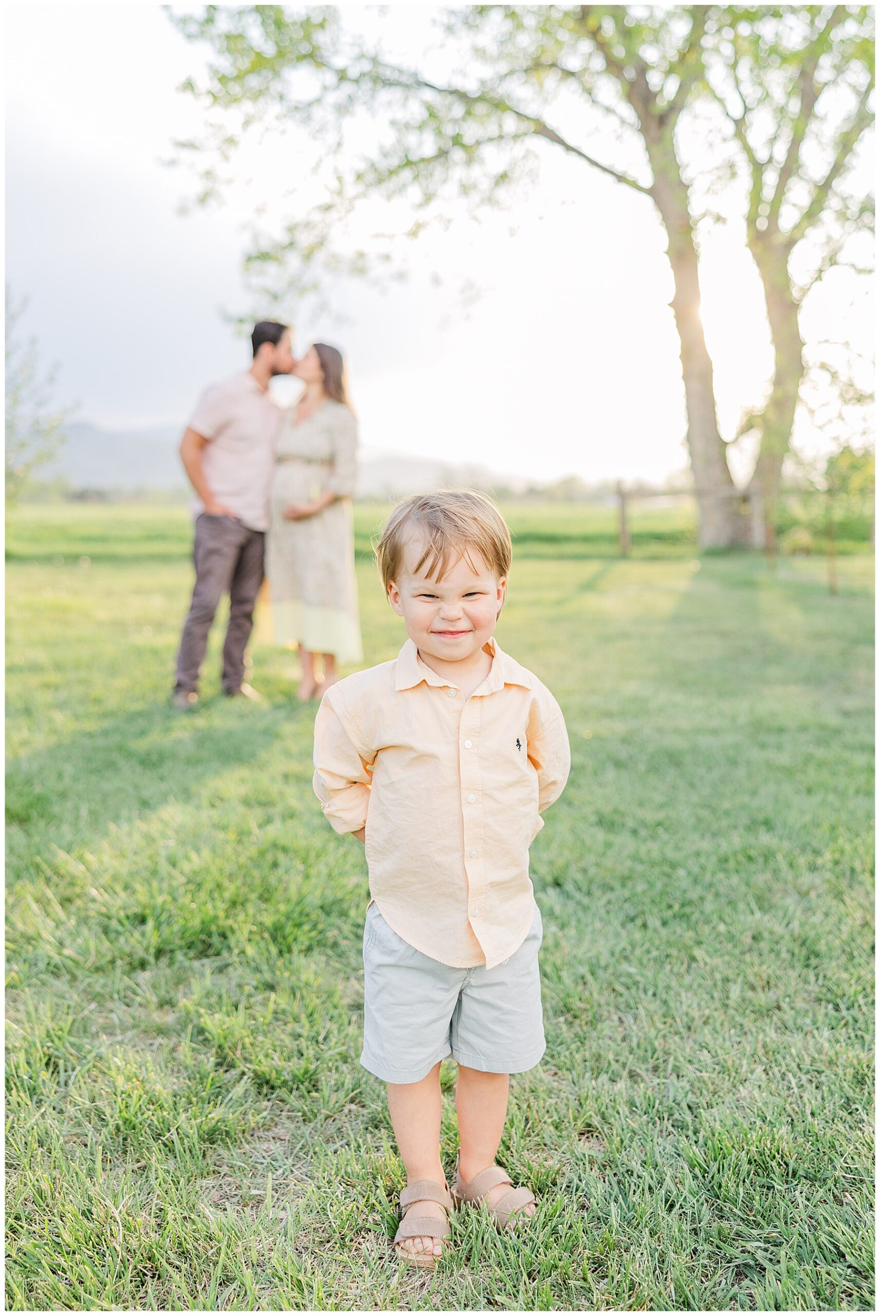 Toddler boy focused at upclose smiles for the camera with expecting parents kissing in the background during Peterson Family Maternity Session | Ya Ya Orchard Longmont, CO