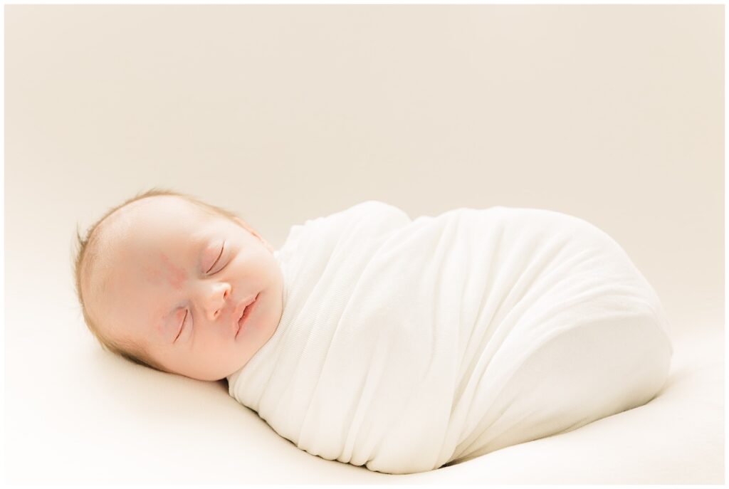 Baby girl swaddled in white with a neutral colored background sleeps during light and airy in-home newborn session with Catherine Chamberlain Photography
