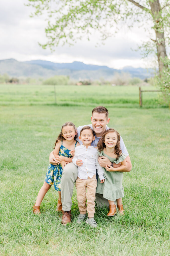 Dad squats behind his three kids and he hugs them during Lantzy Extended Family Session, Longmont, CO