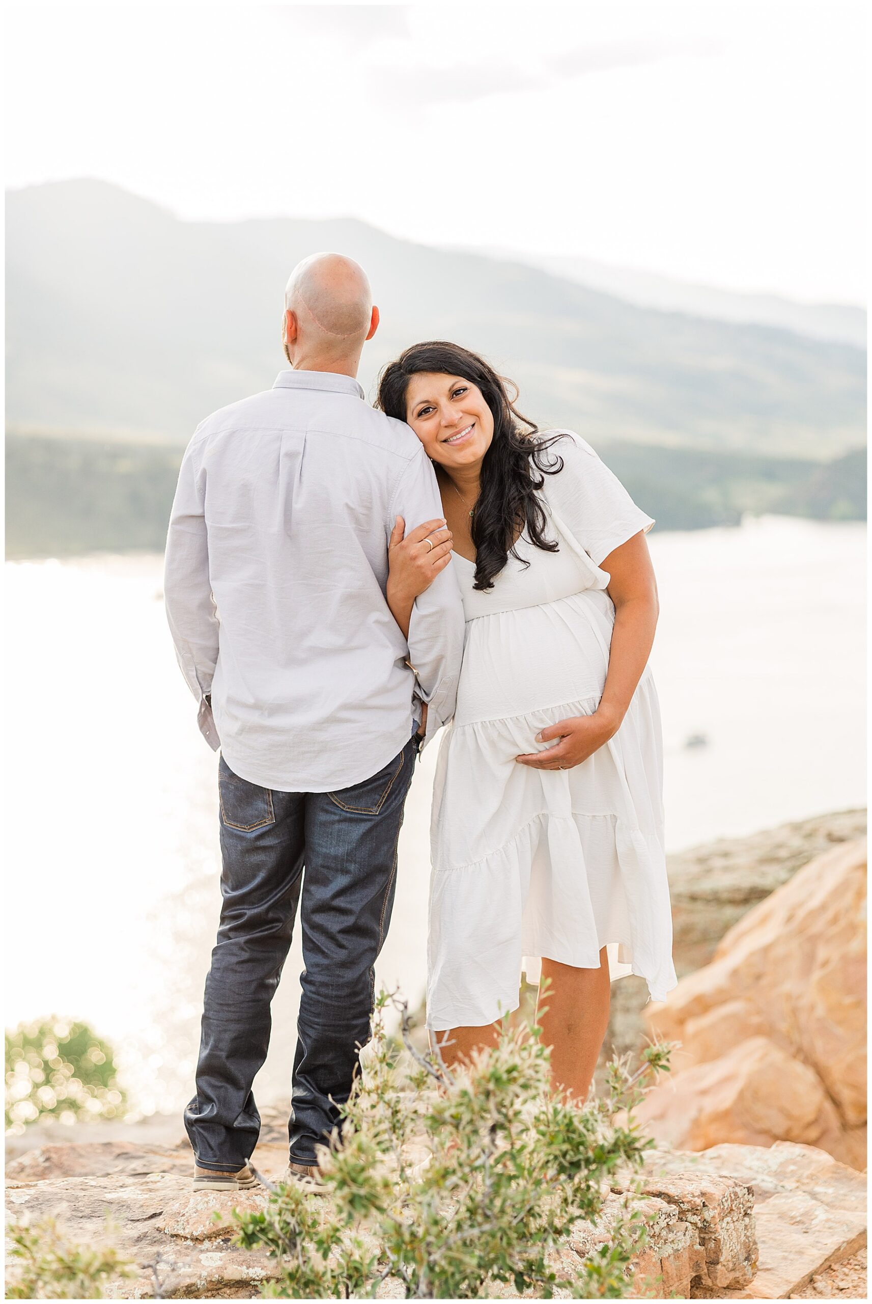 Expecting couple pose as father looks out over a river and wife smiles at the camera for Agnello Family Maternity Session at Horsetooth Reservoir, Fort Collins, CO