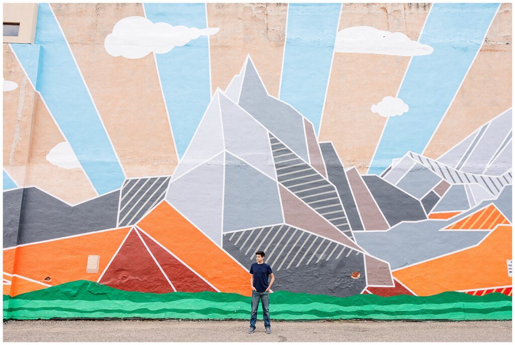 Teen puts his hands in his pockets while standing in front of a mountain mural for Alexander’s Outdoor Senior Session in Fort Collins, CO
