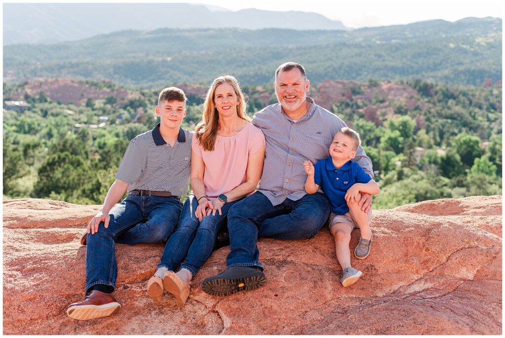 Family of four sit and put their arms around each other with a view of the valley behind them during Dixon Outdoor Family Session at Garden of the Gods in Colorado Springs, CO