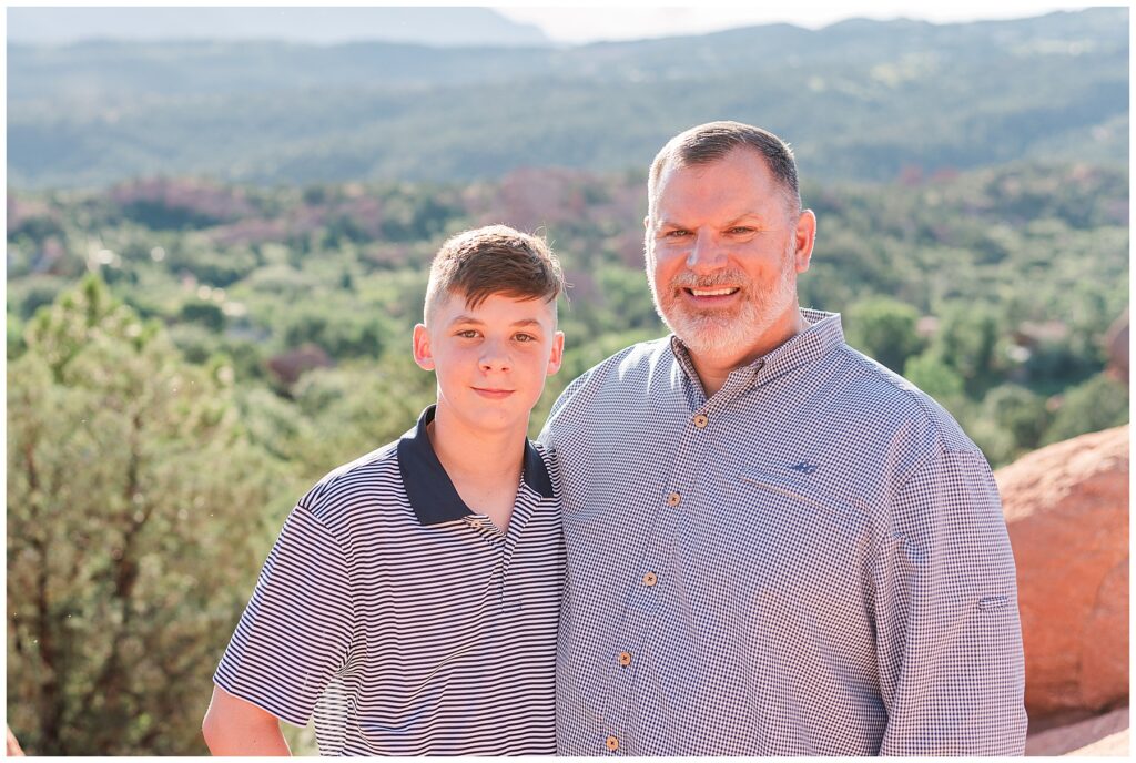 Dad and his eldest son stand with and arm around each other with a large mountain range behind them during the Dixon Outdoor Family Session at Garden of the Gods in Colorado Springs, CO
