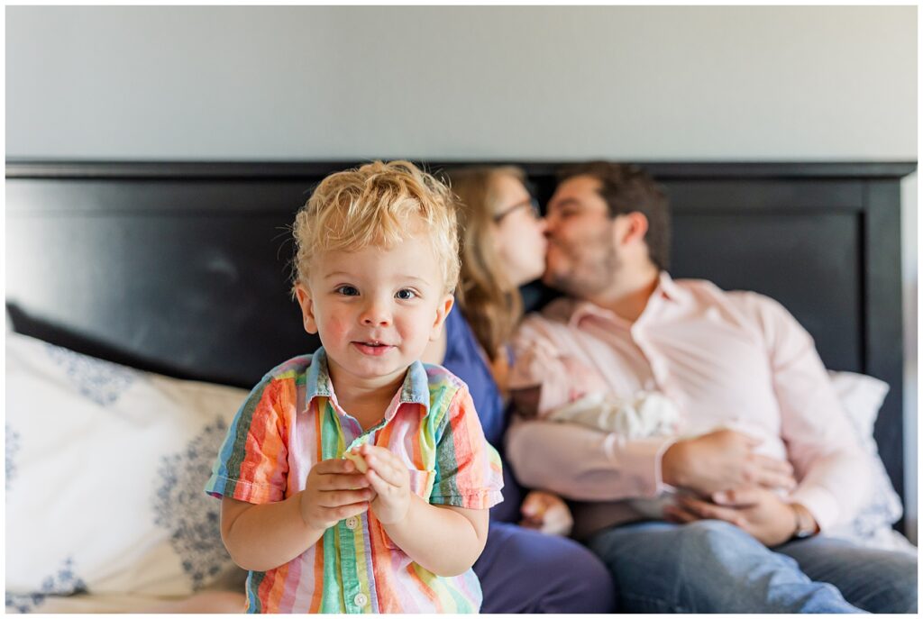 Young toddler boy poses for a close up with his parents and newborn baby sister behind him for the Schwartz family newborn session