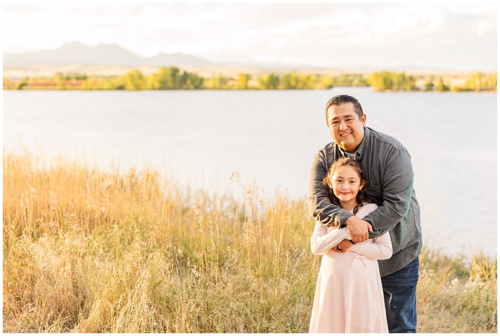 Dad hugs his daughter in front of a lake during the Romero Family Fall Session at Standley Lake in Westminster, CO