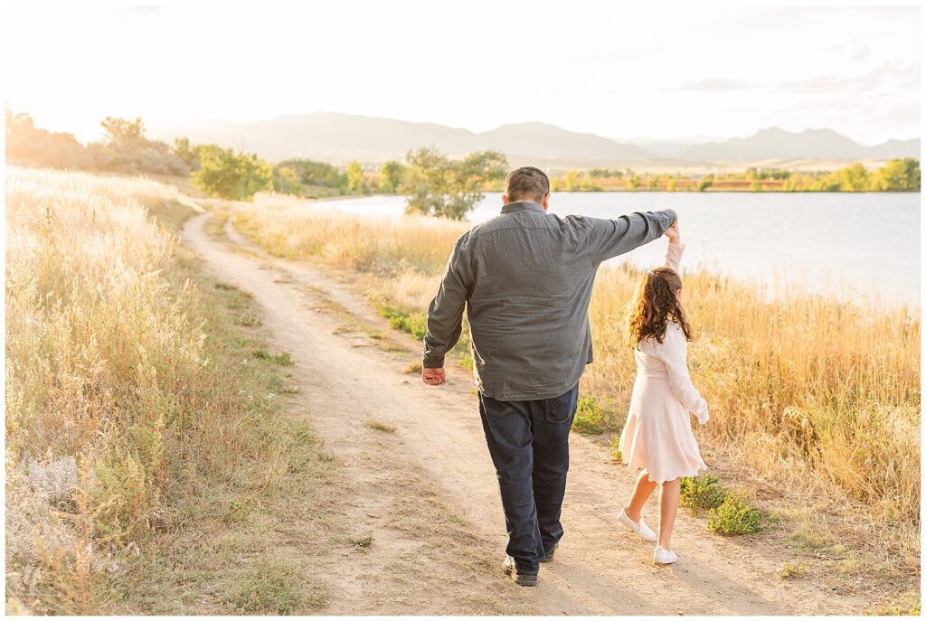 Father and daughter walk along a trail and he holds out his arm to twirl his daughter  and they walk toward the mountain