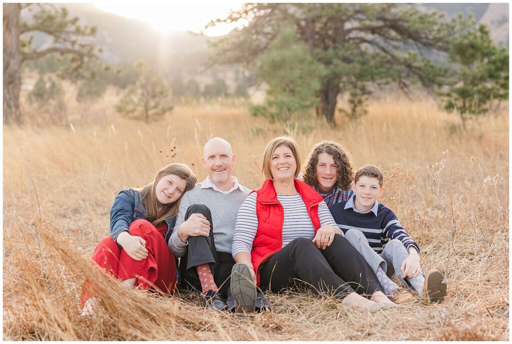 Family of five all sit on the ground in tall golden grass and look at the camera smiling