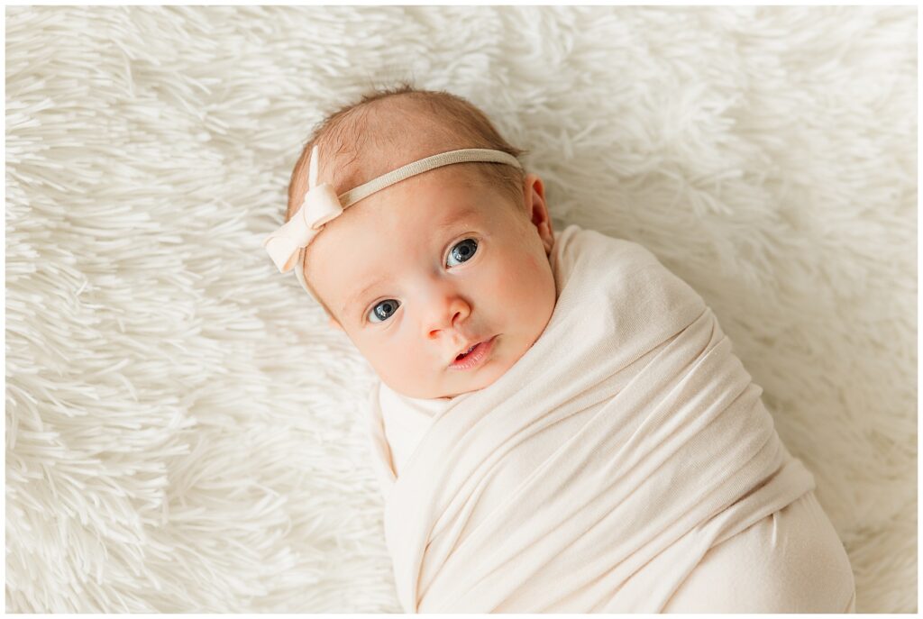 Little newborn baby poses in a swaddle with a bow on and eyes open