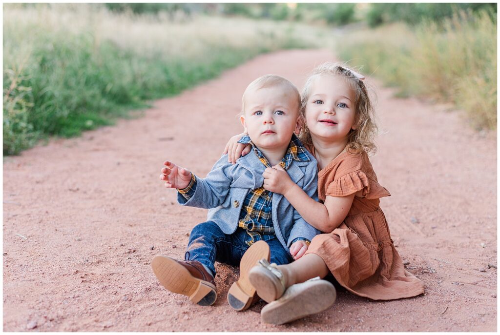 Toddler brother and sister sit together on a dirt pathway