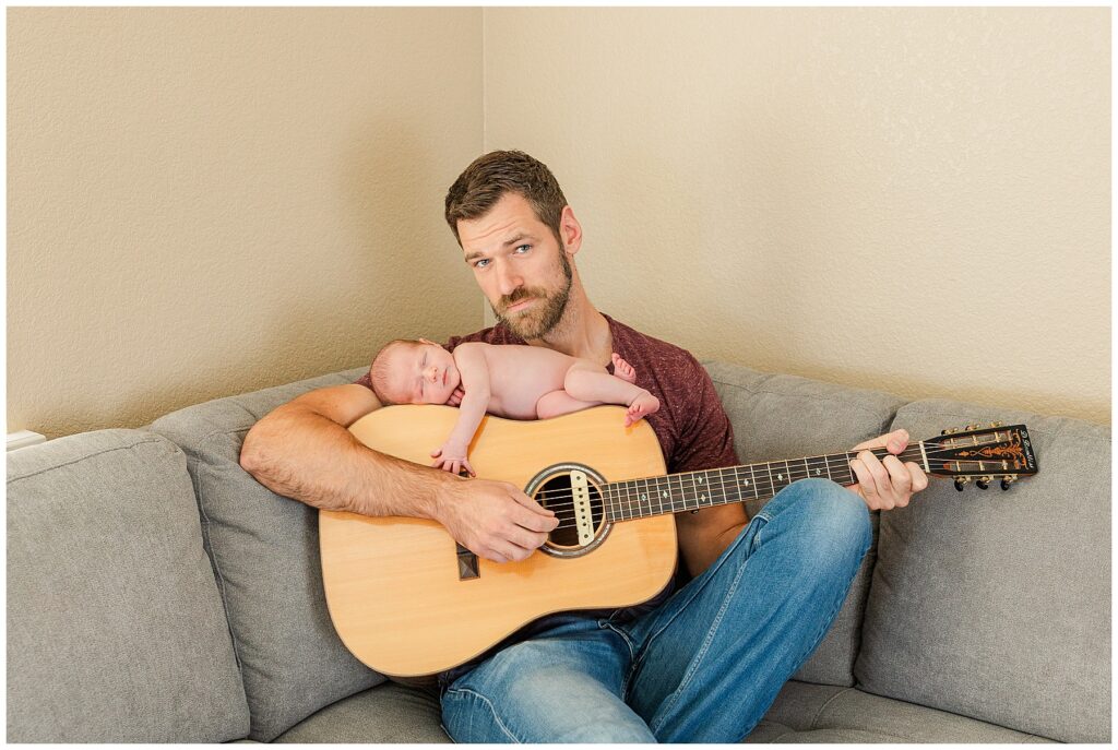Dad sits on the couch with his newborn baby on top of his guitar