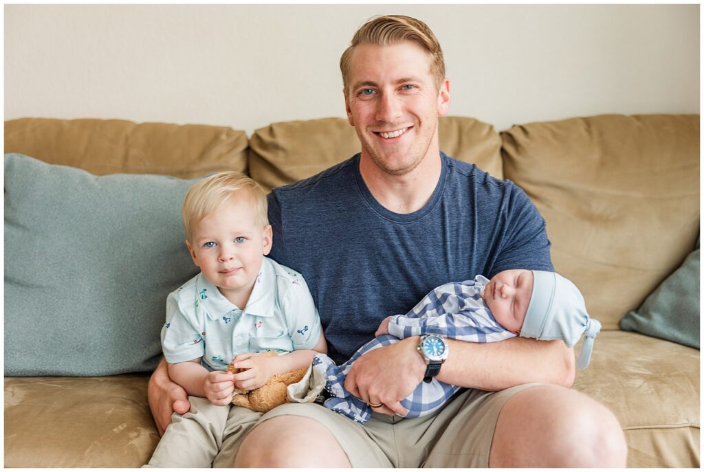 Dad sits on the couch with his toddler boy and newborn baby