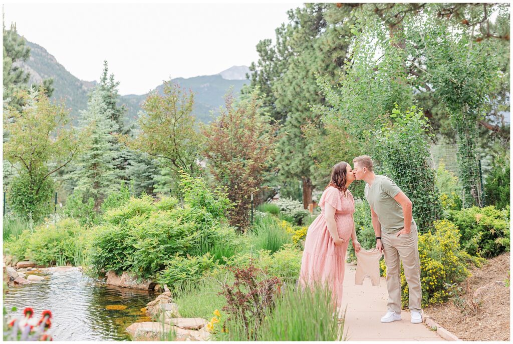 Expecting couple share a kiss while hold a baby outfit between them